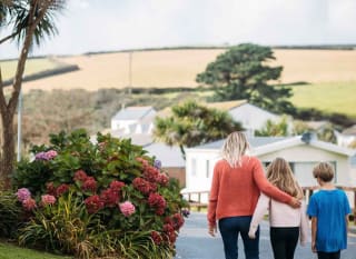 A woman with blonde hair wearing an orange jumper and jeans has her arm around a girl with light brown hair, who is wearing a pale pink top and black trousers. They are walking together down a path at our holiday park, Newquay Bay, Cornwall. image