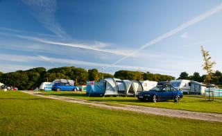 A row of pitched tents, caravans and cars are parked up on a grass parking spot on a sunny day. image