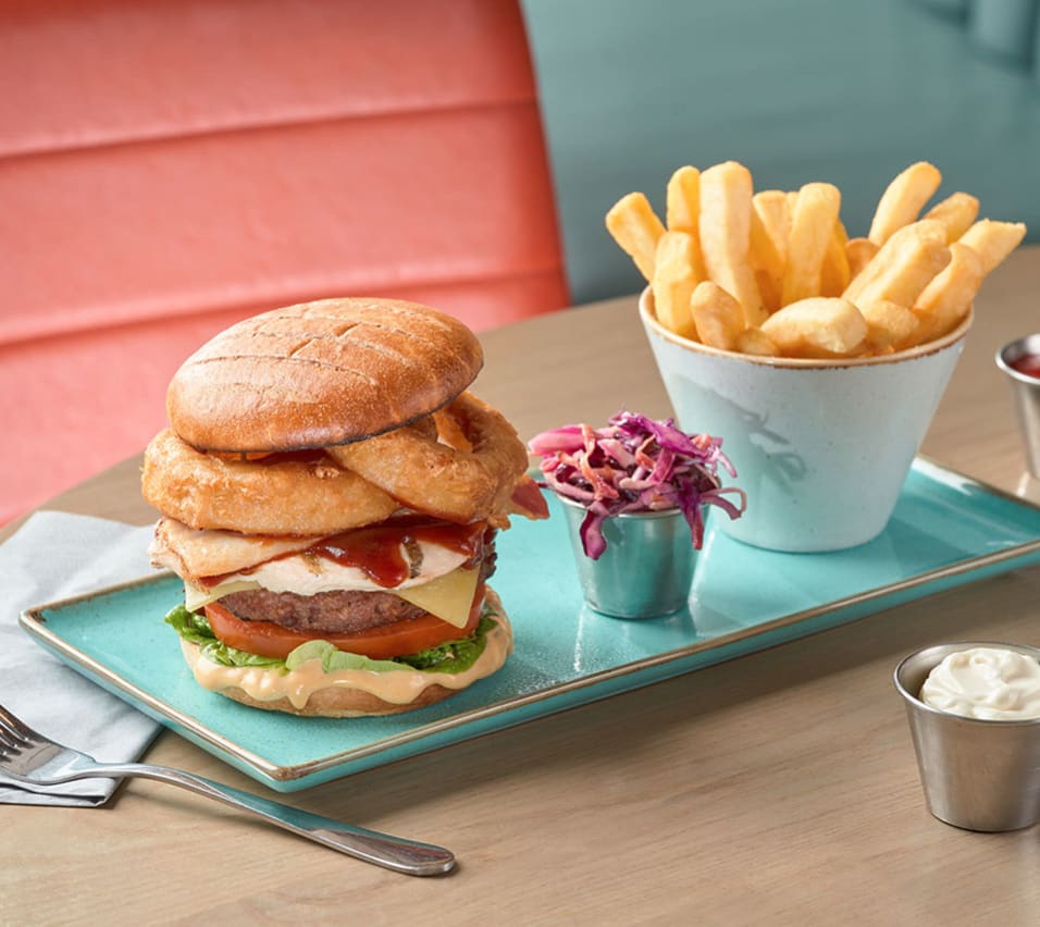 A rectangle plate with a white ceramic holder of chips, a tall stacked burger and a small metail container with red cabbage. image