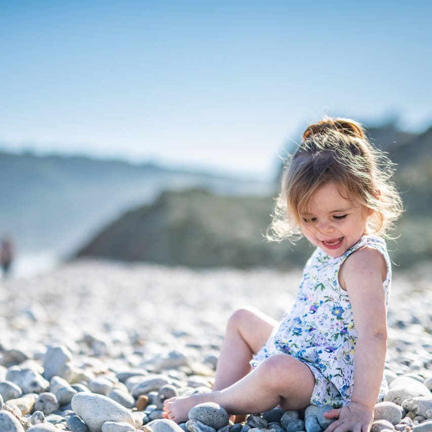 A young girl sitting on a pebble beach wearing a light blue flowered dress on a hot sunny day near one of our holiday parks.