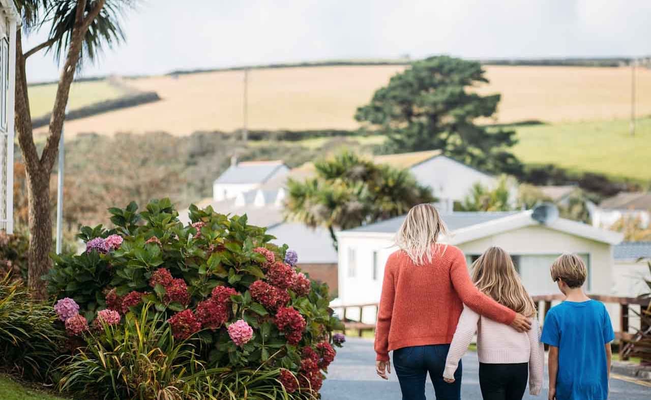 A woman with blonde hair wearing an orange jumper and jeans has her arm around a girl with light brown hair, who is wearing a pale pink top and black trousers. They are walking together down a path at our holiday park, Newquay Bay, Cornwall. image