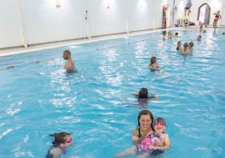 Lots of people of all age ranges swimming in an indoor swimming pool. There is a lifeguard sitting at the site of the pool on a lookout chair.