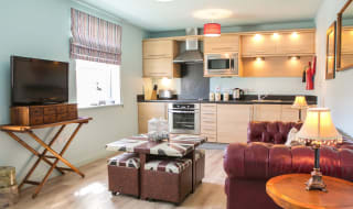 An open-plan view of one of our apartments. A wooden modern kitchen spreads across the back wall, with a red leather sofa on the right side, a compact four-seater table in the middle with a TV opposite the sofa. image