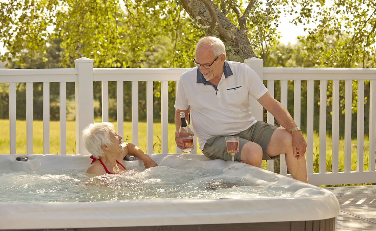 A couple lounging in the hot tub at The Lakes Rookley. The female is sat in the hot tub, whilst the male is sat alongside the hot tub. They are enjoying a bottle of wine together. image
