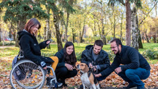 two women and two men are pictured with a medium sized dog. One woman is in a wheelchair and the others are crouched down petting the dog.