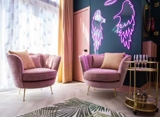 The stunning lounge area in The Esme Compact. There are two plush velvet chairs, a gold drinks trolley and some pink neon lighting in the shape of angel wings. image