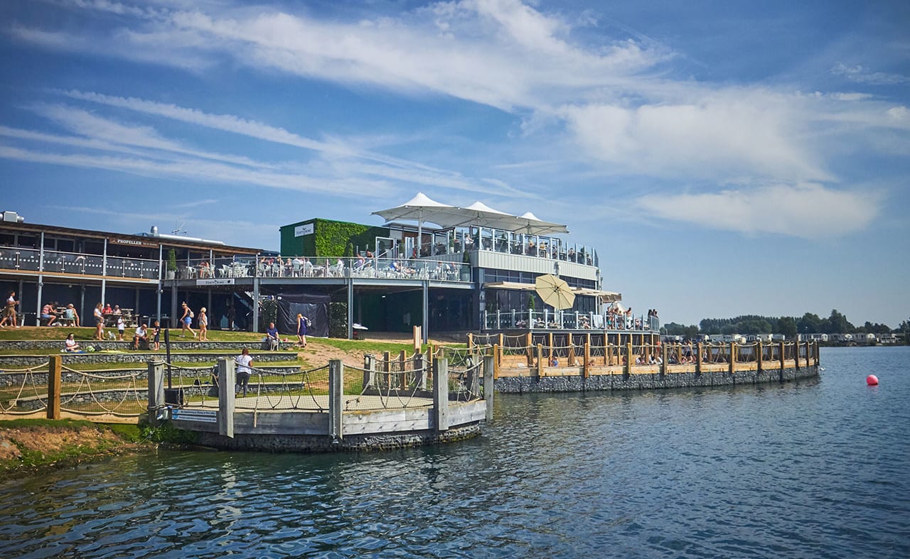 A photo of a restaraunt with a ground floor and two balconey levels of outdoor steating. There are lots of people sitting across all levels and the building is next to a large river. image