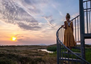 A photo of a girl with blonde hair in a bun wearing a orange polkadot dress, whom is standing in the middle of a spiral staircase looking out at the sunset.