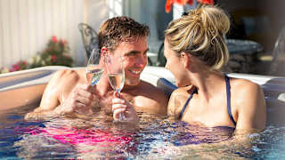 A happy couple smiling in a hot tub on a sunny day. They are doing a cheers with champagne glasses. The man has short brown hair and is shirtless with pink swim shorts on. The woman has blonde hair and is wearing a blue swimsuit.