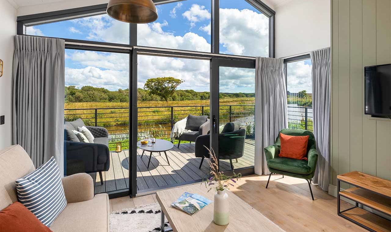 An interior view of inside a living room of a barn. The windows meets the celing with light grey curtains, there is a wooden table and fabric covered dark grey chairs on a wooden patio with the sun shining on them. image