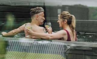 A middle-aged couple are cuddled with drinks in hand, whilst sitting in a hot tub. image