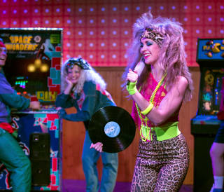Two women are dressed up in 80's party attire with big curly hair inside an 80's themed disco with an arcade machine.
