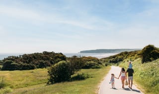 A young family are walking along a dry dirt path which is situated between rolling green hills on a hot summer's day. image