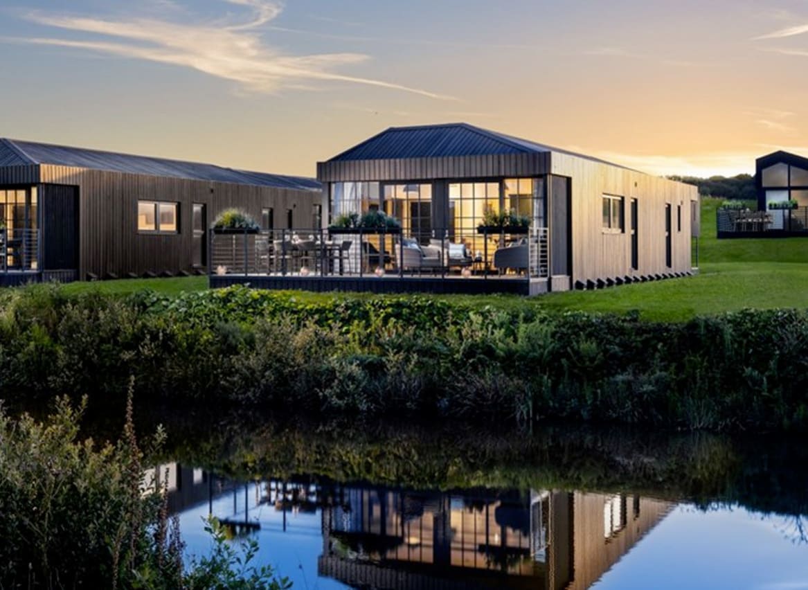 One of our holiday home barns. The sun is setting behind the spacious barn exterior, which sits on the green grass near the lake. The lights are on inside. image