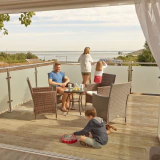 A family of four on the balcony of their holiday home in The Bay Colwell. There are two adults, a young boy, and a young girl with strawberry blonde hair, and all dressed in summer attire. image