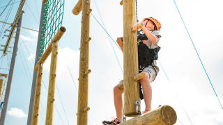 A pre-teen child is climbing on a wodden structure with a bright orange helmet, torso harness and a pully rope system. The child is looking up to judge his next move as he accends higher on the structure.