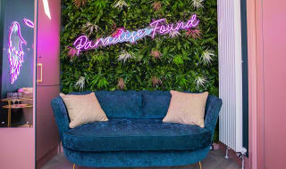 A uniquely decorated seating area with fake vines on the wall, a blue loveseat and a pink neon sign which says, ''Paradise Found