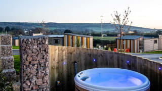 The outside of one of our holiday parks, which has an empty hot-tub behind a private fence. The sun is setting behind the wide view of the holiday park. image