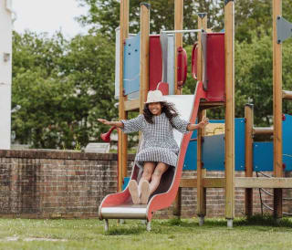 A wooden children's adventure playground outside on some trimmed grass at Newquay Bay, Cornwall. A girl wearing a white hat and a checkered dress is sliding down a red slide.