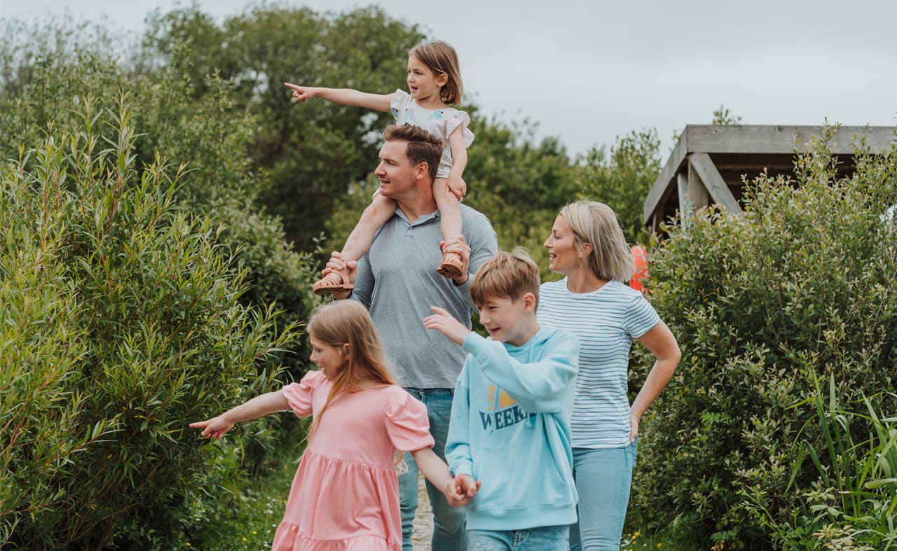 A family of five strolling through one of our holiday parks. The man is carrying the young girl on his shoulders, and the other young boy and girl are walking. The family is pointing at something in the distance. the surrounding area is green. image
