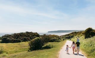 A young family are walking along a dry dirt path which is situated between rolling green hills on a hot summer's day. image