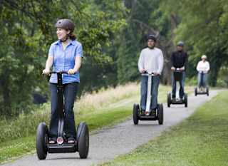 A group of middle-aged adults of multiple races are segwaying down a pathway, one behind the other. On either side of them are grass and tree areas. image
