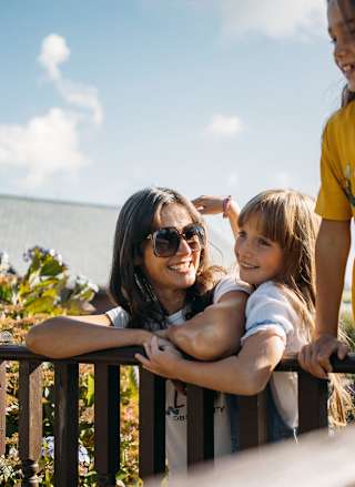 A family enjoying their stay at one of our holiday parks. There is an adult woman and two young girls. One of the girls is hugging the woman, the other is leaning over a banister. All three have wide smiles.
