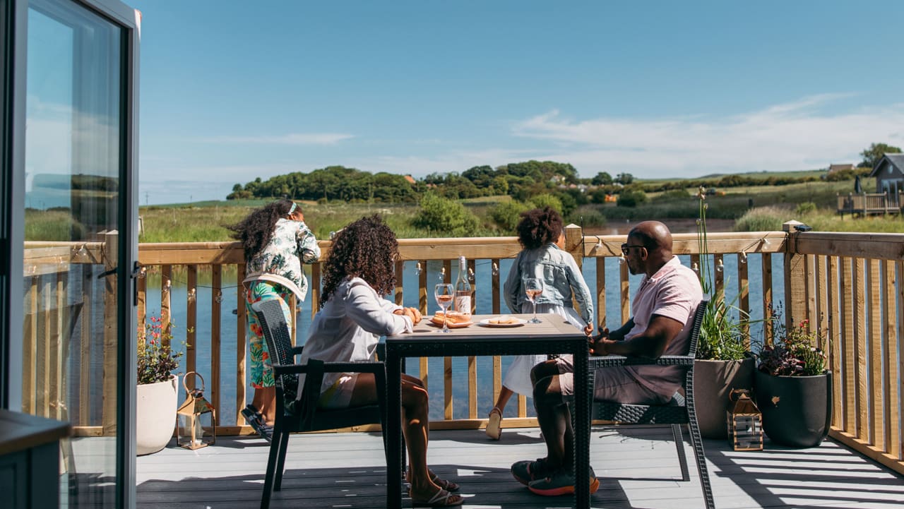 A family sat outside on the decking of their holiday home on a sunny day at The Bay Filey, Yorkshire, overlooking the lake and woodland views.
