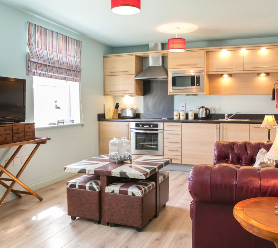 An open-plan view of one of our apartments. A wooden modern kitchen spreads across the back wall, with a red leather sofa on the right side, a compact four-seater table in the middle with a TV opposite the sofa. image