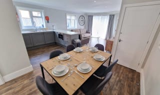 An open-plan view of one of our cottages. In front view is a dinner table set for six. across from the table is a U-shaped Kitchen area with plenty of worktop space. In the background is the living room area containing two sofas. image