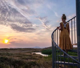 A photo of a girl with blonde hair in a bun wearing a orange polkadot dress, whom is standing in the middle of a spiral staircase looking out at the sunset.