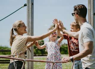 A young family are all balancing on a wooden beam with a rope handhold engaging in a group high five. image