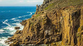 A landscape image of the sea and cliffs located at Lands End. A blue sky with minimal clouds sets the scene with the Land's end tourist building seen in the top right of the image.