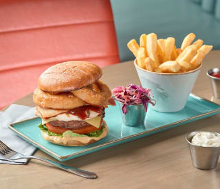 A rectangle plate with a white ceramic holder of chips, a tall stacked burger and a small metail container with red cabbage.
