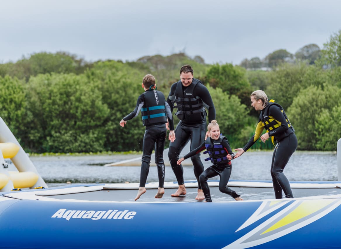 A young family wearing life jackets and full wetsuits balancing on a inflateable in a body of water at one of our holiday parks. image