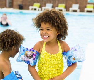 Two young children paddling in an outdoor pool. One is wearing a bright yellow swimming costume with armbands and the other is mostly out of shot but some of their curly hair is in frame.