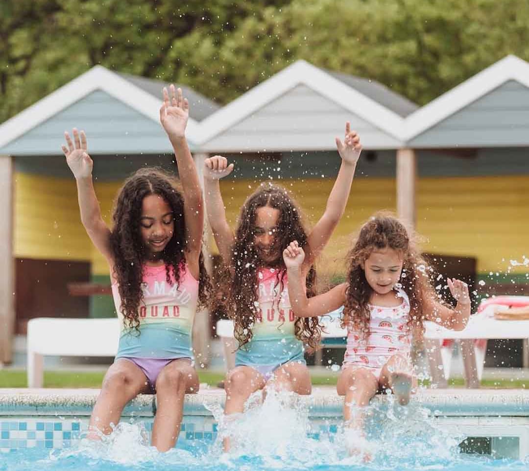Three girls, each with long brown curly hair and pink swimsuits, are splashing and paddaling on the edge of our outdoor pool at Newquay Bay, Cornwall image