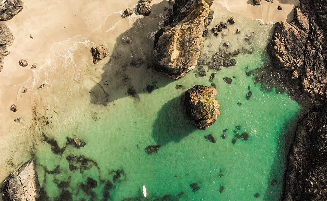 A drone image of Kynance Cove, Cornwall. There are lots of rocks to explore, a sandy coastline and lovely blue waters. image