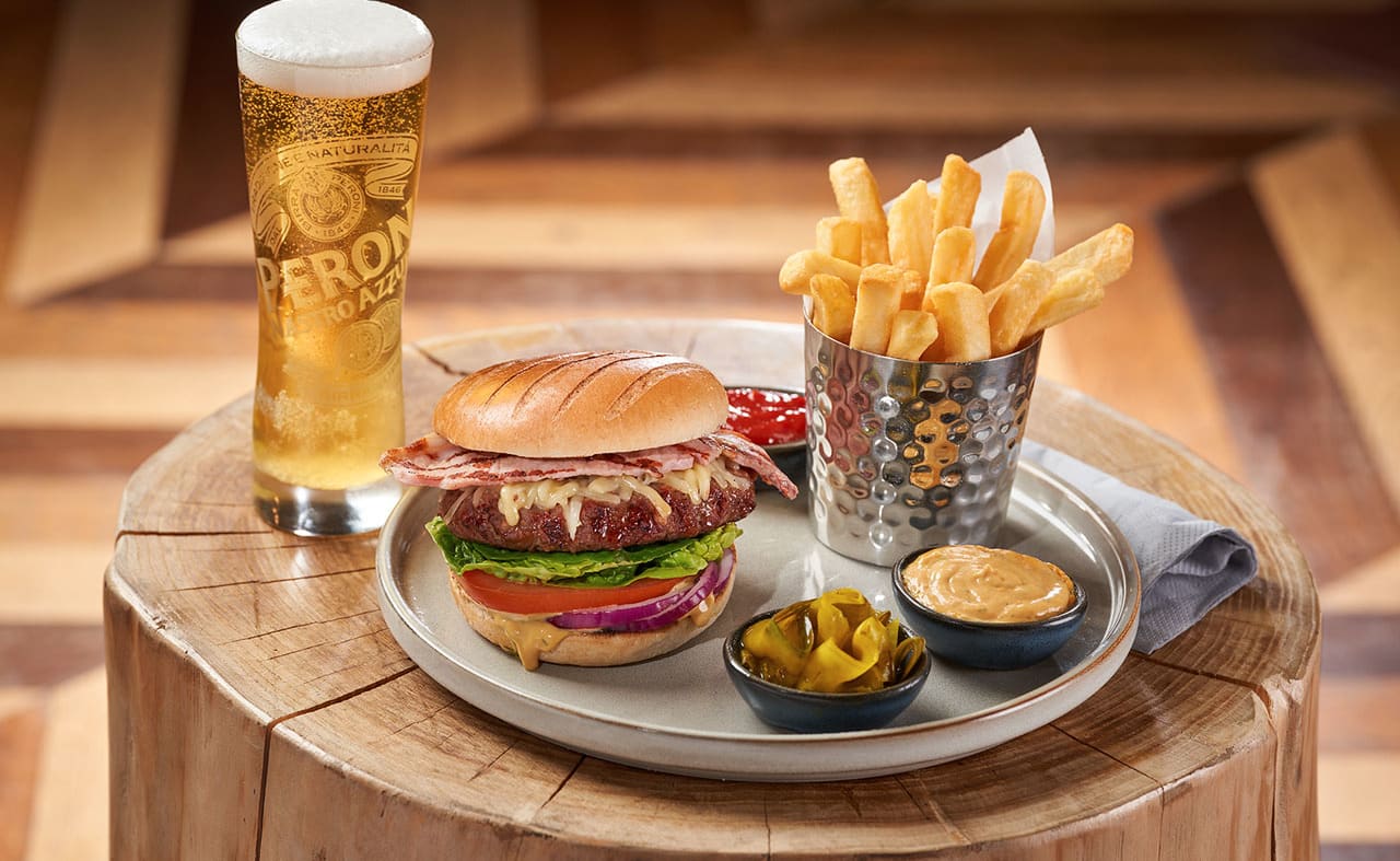 On the place is a stacked bacon cheeseburger with lettace, tomato and sliced red onion. Next to the burger is cooked chips and ketchup, pickles and burger sauce. Next to the plate on the left is a tall glass of larger with a thick, creamy head. image