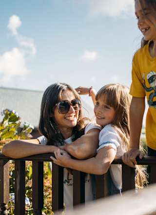 A family enjoying their stay at one of our holiday parks. There is an adult woman and two young girls. One of the girls is hugging the woman, the other is leaning over a banister. All three have wide smiles.