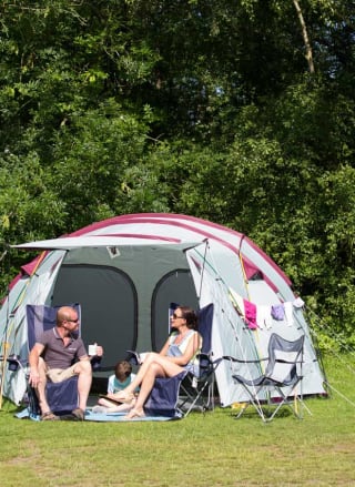 A pitched up family tent with a man, woman and baby sitting in the porch area of the tent. A teen boy and girl are cycling by on a hot sunny day.