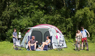 A pitched up family tent with a man, woman and baby sitting in the porch area of the tent. A teen boy and girl are cycling by on a hot sunny day. image