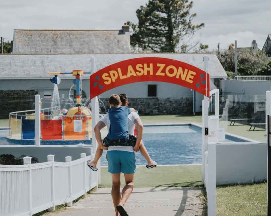 A young boy on his father's back heading towards the outside pool area. A read sign with yellow writing reads 'Splash Zone'.