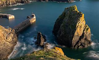 An image of Mullion Cove, Cornwall on a sunny  day. There are views of the vast cliffs, rocks and stunning blue sea. image