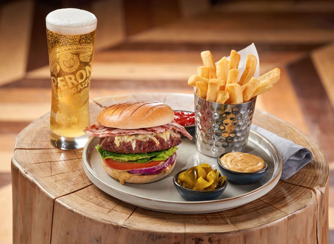 On the place is a stacked bacon cheeseburger with lettace, tomato and sliced red onion. Next to the burger is cooked chips and ketchup, pickles and burger sauce. Next to the plate on the left is a tall glass of larger with a thick, creamy head. image