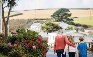 A woman with blonde hair wearing an orange jumper and jeans has her arm around a girl with light brown hair, who is wearing a pale pink top and black trousers. They are walking together down a path at our holiday park, Newquay Bay, Cornwall. image