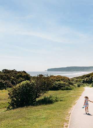 A young family are walking along a dry dirt path which is situated between rolling green hills on a hot summer's day.