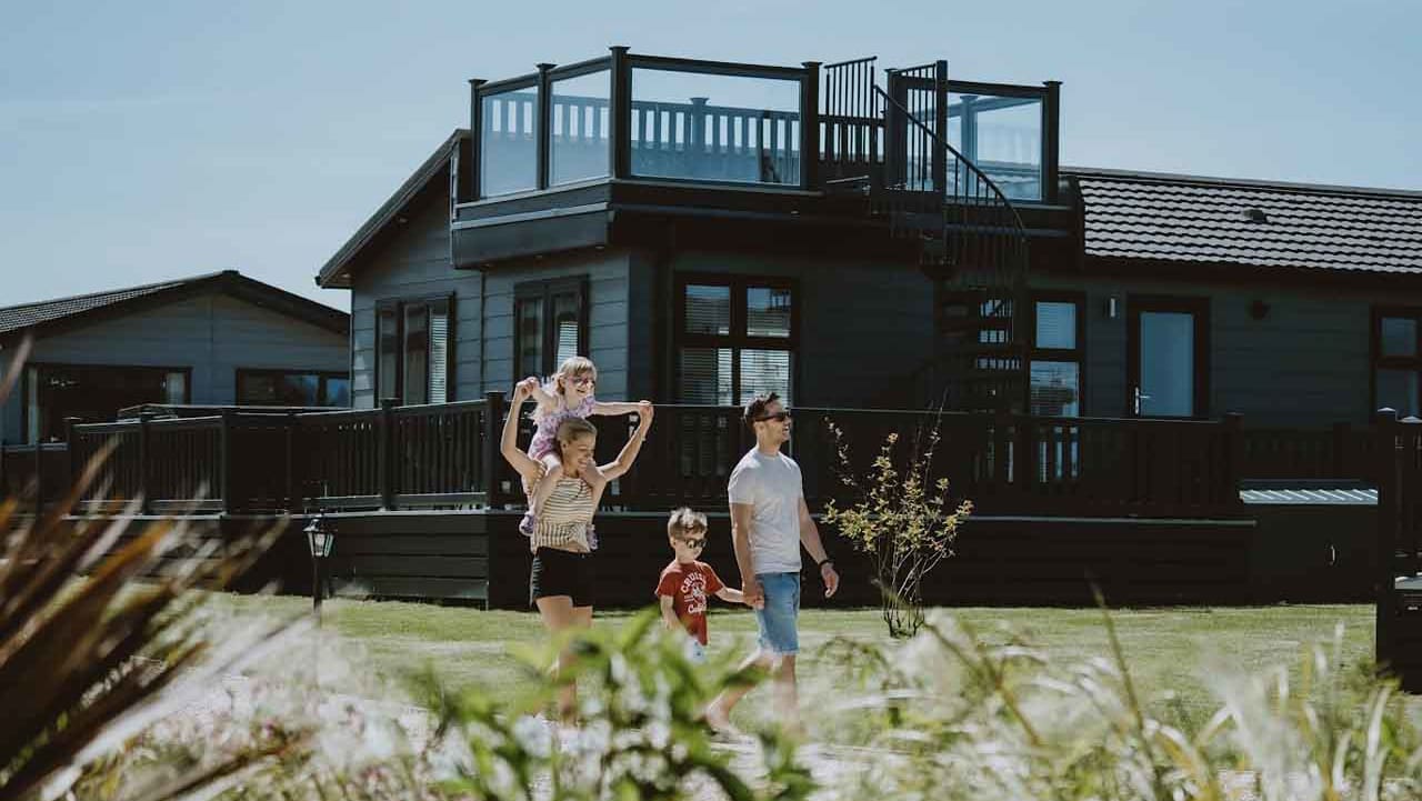 A young family taking a stroll past our stunning holiday homes with roof terraces. One of the two children is on their mum's shoulders.
