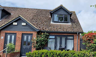 An outside view of one of our 3-bed cottages. Bushes outside lead to three steps to the pathway of the front door to the cottage. The sky above is blue yet very cloudy. image