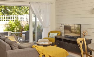 An open-plan view of one of our cottages, with a round table to the right, a brown sofa facing a tv on a unit. Next to the sofa is a set of french doors which are opened up onto a decking area. On the decking is a garden table and chairs next to a hot tub image